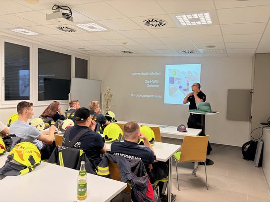 Feuerwehr-Schulung in Österreich: Vortragender erklärt HWS-Schiene vor Publikum mit Helmen.